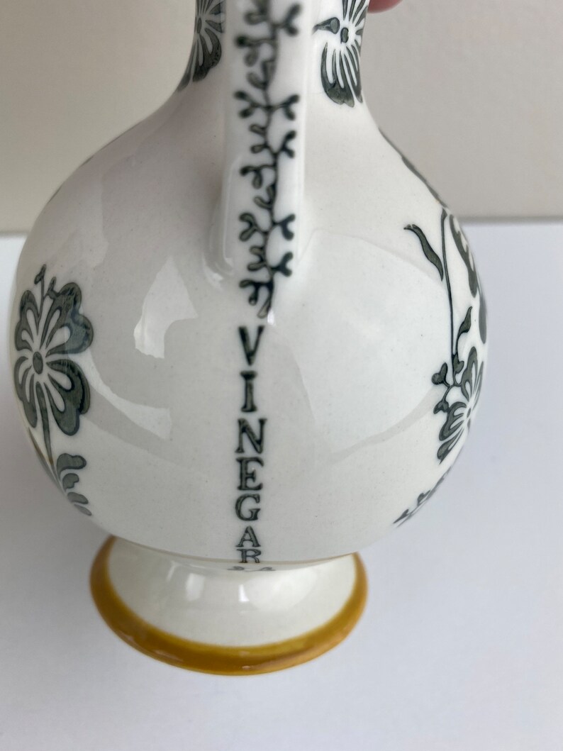 Może przedstawiać: Biała ceramiczna butelka na ocet z czarnym wzorem kwiatowym i napisem "VINEGAR" napisanym pionowo na boku. Butelka ma ż&oacute;łtą obw&oacute;dkę wok&oacute;ł podstawy.