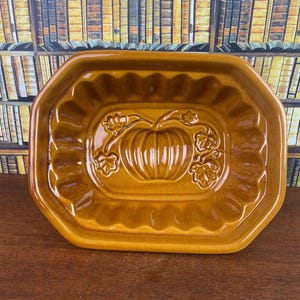May include: A ceramic, octagonal-shaped baking dish in a warm, golden-brown hue. The dish features a raised pumpkin design in the center, surrounded by decorative leaves. The edges have a scalloped design. A vintage kitchenware item.