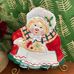 May include: A ceramic Mrs. Claus plate, featuring a red and white outfit and holding cookies. The plate has a green and red plaid scarf and a white bonnet. The plate is displayed on a gold metal stand.