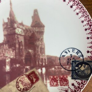 May include: A white ceramic plate with a burgundy border and a vintage design featuring a castle, a bridge, and old postage stamps. The plate has the word "AVIERS" in a blue circle stamp.