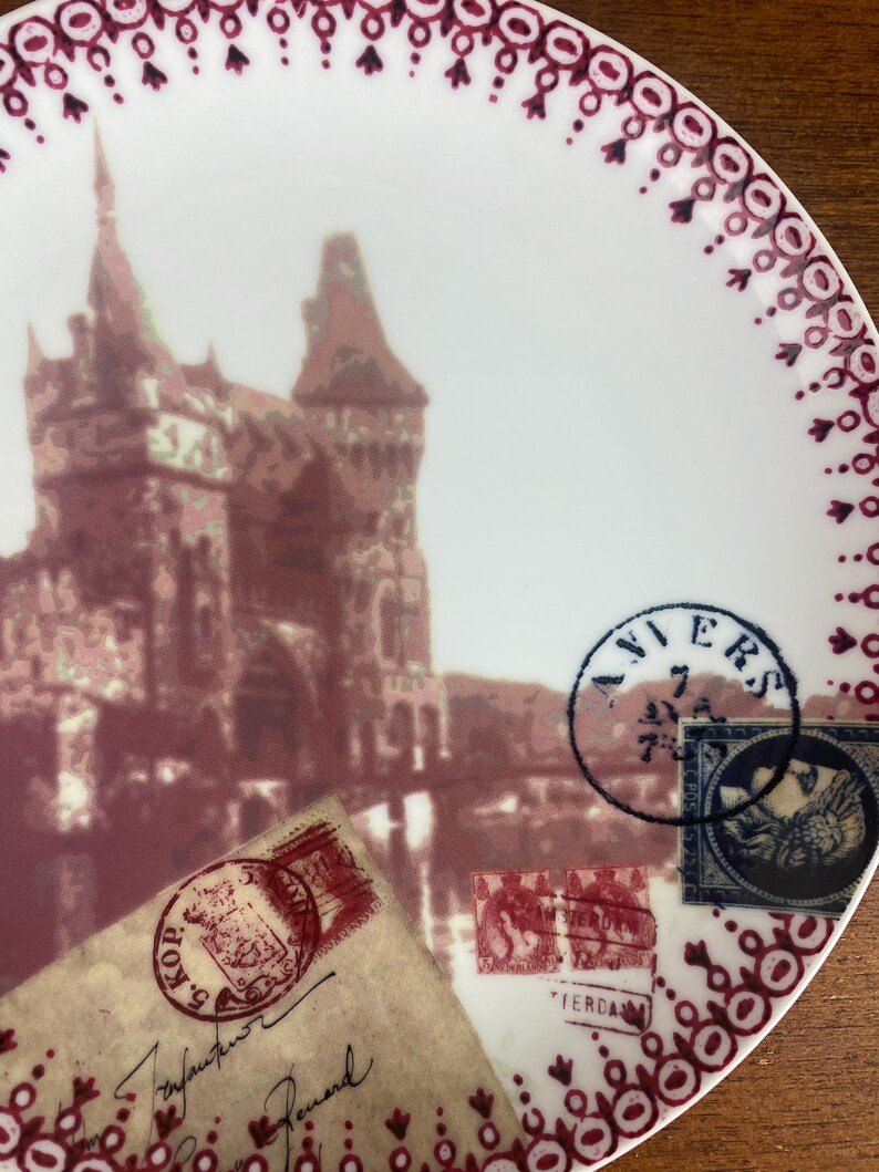 May include: A white ceramic plate with a burgundy border and a vintage design featuring a castle, a bridge, and old postage stamps. The plate has the word "AVIERS" in a blue circle stamp.