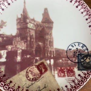 May include: A white ceramic plate with a red and white border. The plate features a vintage image of a castle with a red and white color scheme. The image is surrounded by vintage postage stamps with red and blue accents.