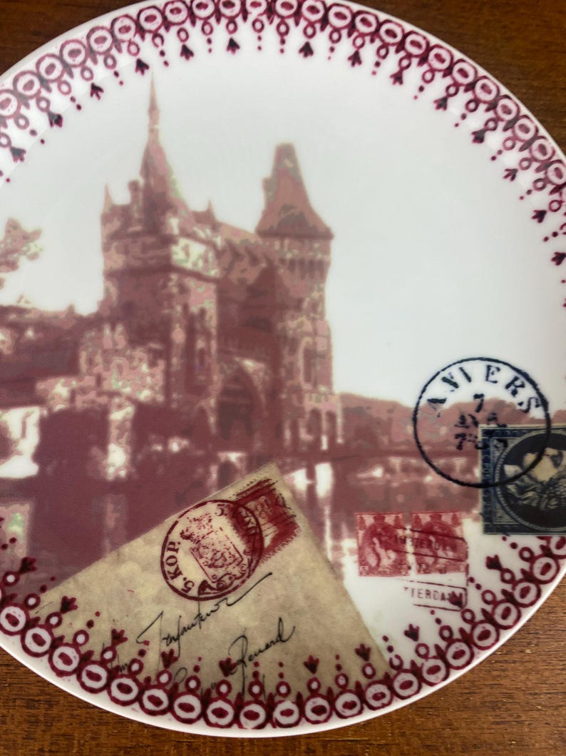 May include: A white ceramic plate with a red and white border. The plate features a vintage image of a castle with a red and white color scheme. The image is surrounded by vintage postage stamps with red and blue accents.