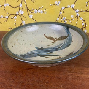 May include: A ceramic bowl with a speckled beige interior and a dark blue rim. The bowl features a blue and brown painted wave design. The background is a yellow floral pattern.