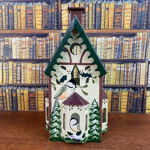 Può includere: Un portacandele decorativo a forma di chiesa dipinto a mano. La struttura bianco sporco presenta un tetto e finiture verdi, con illustrazioni di uccelli e agrifoglio. Il portacandele è alto circa 20 cm.