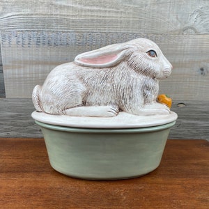 May include: A white ceramic rabbit figurine sits atop a green ceramic serving dish. The rabbit has pink ears and is holding a small orange carrot.