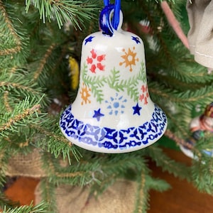 May include: A white ceramic bell Christmas ornament with a blue ribbon. The bell is decorated with red, orange, and blue floral patterns and green leaves. The bottom edge has a blue and white geometric design. The ornament hangs on a green Christmas tree.