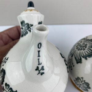 Może przedstawiać: Zestaw ceramicznych butelek na olej i ocet w kolorze białym z czarnymi wzorami kwiatowymi i złotymi akcentami. Butelka na olej ma napis "OIL" napisany pionowo na boku.