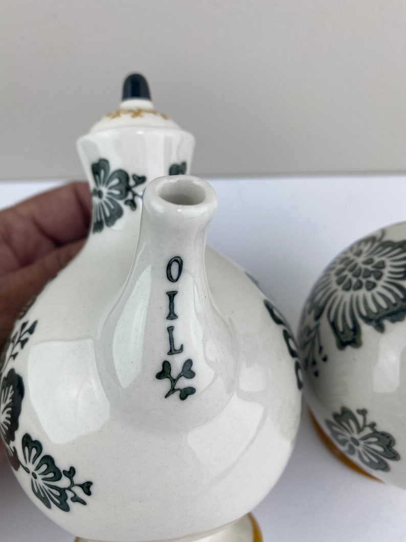 Może przedstawiać: Zestaw ceramicznych butelek na olej i ocet w kolorze białym z czarnymi wzorami kwiatowymi i złotymi akcentami. Butelka na olej ma napis "OIL" napisany pionowo na boku.