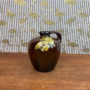 May include: A dark brown ceramic jug with a handle, featuring a green and yellow leaf and berry motif. The jug has a glossy finish and is displayed on a wooden surface.