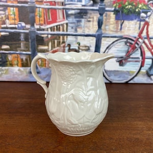 May include: A white ceramic pitcher with a handle and a scalloped rim. The pitcher features embossed floral and leaf designs. The background includes a painting of a canal scene with a red bicycle.