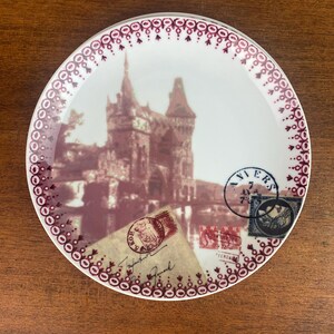May include: A white ceramic plate with a burgundy border and a vintage image of a castle with postage stamps and a handwritten note. The plate has a decorative border of burgundy circles and dots.