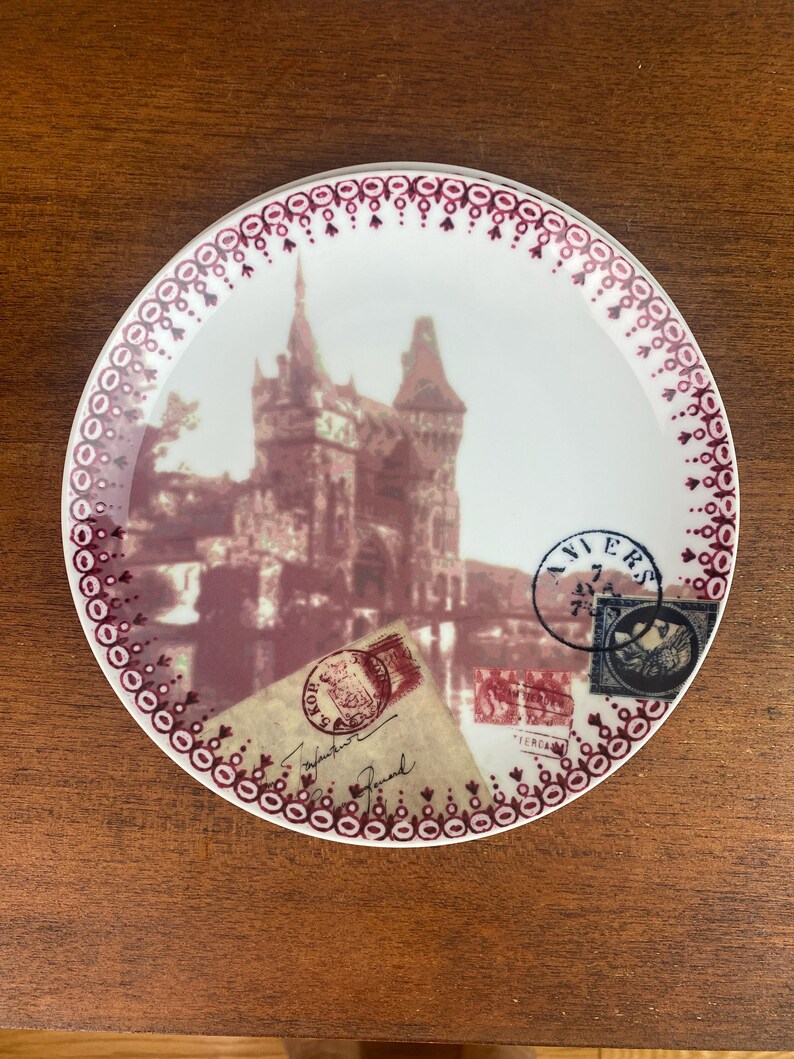 May include: A white ceramic plate with a burgundy border and a vintage image of a castle with postage stamps and a handwritten note. The plate has a decorative border of burgundy circles and dots.