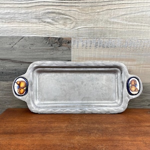 May include: Rectangular metal serving tray with decorative handles. The tray has a textured, silver finish and scalloped edges. Each handle features a blue and white ceramic design with yellow and brown fruit.