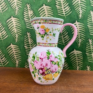 Op de afbeelding: Een witte keramische kan met een roze handvat en bloemenpatroon. De kan heeft roze rozen, gele bloemen en groene bladeren. De rand en de basis hebben decoratieve randen. De achtergrond is groen behang met een varenpatroon.