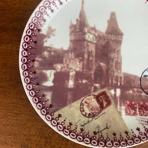 May include: A white ceramic plate with a burgundy border featuring a vintage image of a building with a red roof and a postage stamp with the text "KOP 5" on it.