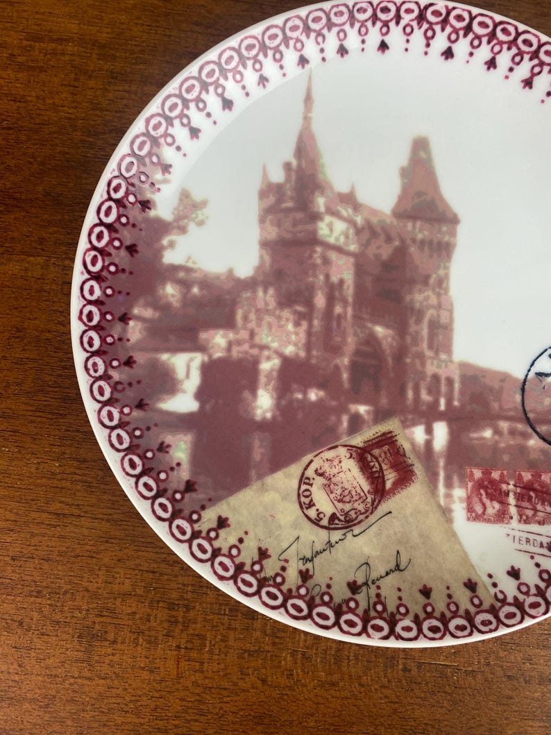 May include: A white ceramic plate with a burgundy border featuring a vintage image of a building with a red roof and a postage stamp with the text "KOP 5" on it.