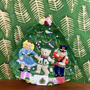 May include: A ceramic Christmas tree ornament with a green base and festive decorations. The ornament features a girl in a blue dress, a teddy bear, and a toy soldier. Santa Claus is at the top of the tree.