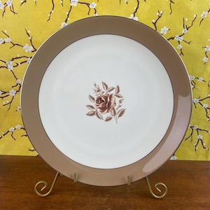 May include: A vintage white plate with a brown border and a gold rim. The plate features a brown rose floral design in the center. The plate is displayed on a gold metal stand, showcasing its elegant design.