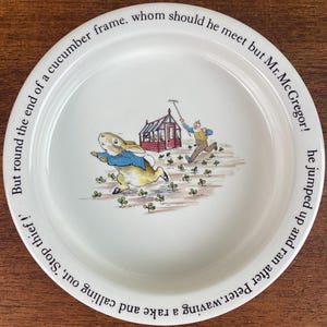 May include: A white ceramic plate with a Peter Rabbit illustration. The plate features Peter Rabbit running away from Mr. McGregor, who is chasing him with a rake. The text around the edge of the plate reads: "But round the end of a cucumber frame, whom should he meet but Mr. McGregor! he jumped up and ran after Peter, waving a rake and calling out, 'Stop thief!'".