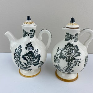 Może przedstawiać: Zestaw dw&oacute;ch ceramicznych naczyń do oliwy i octu z czarnymi i złotymi wzorami kwiatowymi. Naczynia mają białe tło z czarnymi wzorami kwiatowymi i złotymi akcentami.