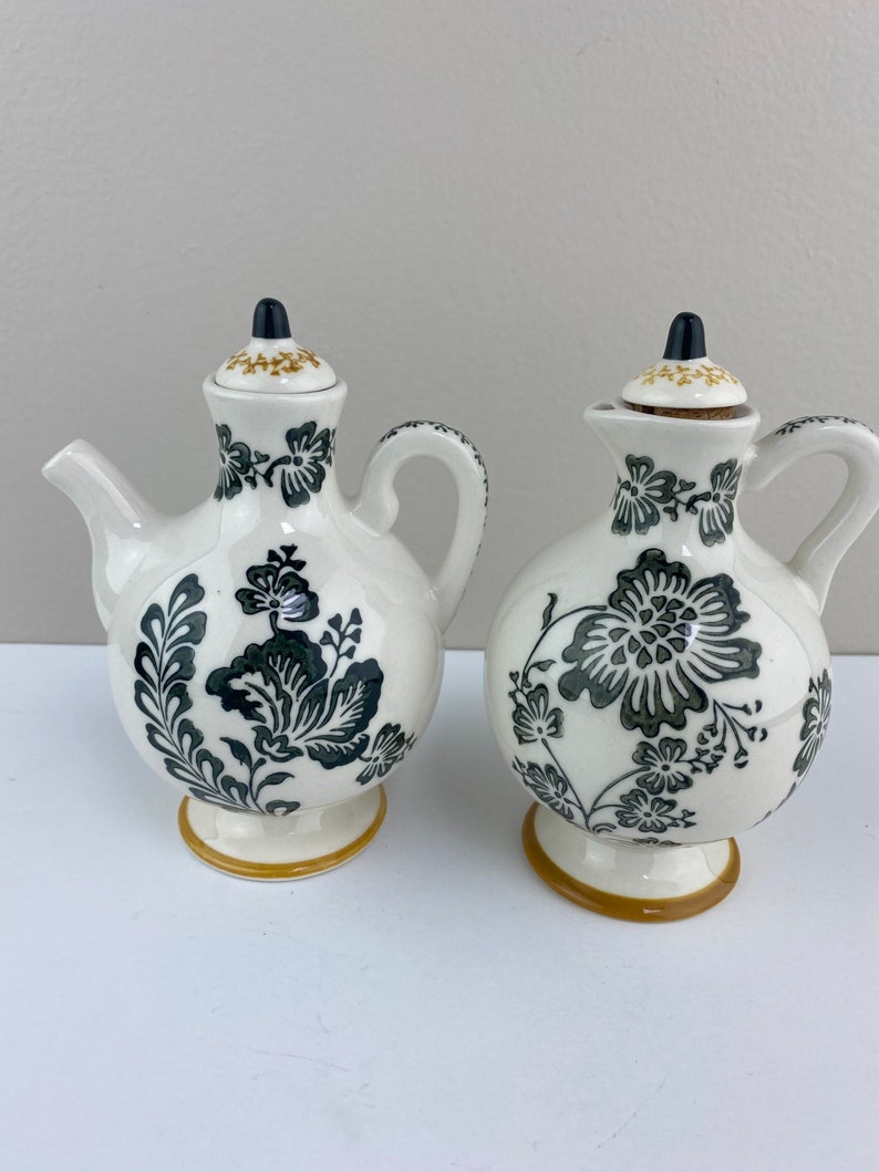 Może przedstawiać: Zestaw dw&oacute;ch ceramicznych naczyń do oliwy i octu z czarnymi i złotymi wzorami kwiatowymi. Naczynia mają białe tło z czarnymi wzorami kwiatowymi i złotymi akcentami.