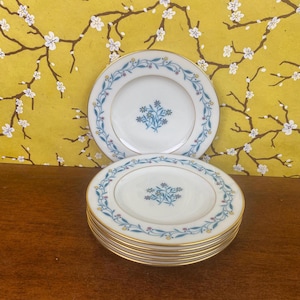 May include: A stack of white porcelain plates with a gold rim and a floral design. The plates have a blue floral border with a central blue floral bouquet. The plates are stacked on a wooden surface.