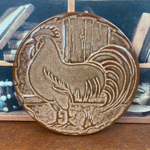 May include: A circular, brown ceramic coaster with a detailed relief of a rooster. The rooster is shown in profile, with its head turned slightly, and is set against a background of vertical lines. The coaster has a rustic, vintage aesthetic.