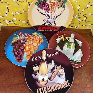 May include: Four round decorative plates featuring wine-themed illustrations. One plate shows two figures holding wine bottles with the text "LES VINS SELECTIONNES AMORCE DANS VOTRE VERRE". Others display grapes, a "Vilforte" wine bottle, and "UN VIN BLANC EXTRAORDINAIRE" with a bottle, bread, and glasses.