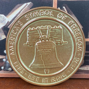 Puede incluir: Un plato redondo de cerámica marrón con la Campana de la Libertad en el centro. El plato está inscrito con "AMERICA'S SYMBOL OF FREEDOM 1776 LET IT RING 1976" en un patrón circular. El plato tiene un acabado brillante.