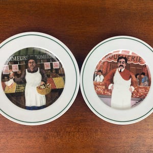 May include: Two white ceramic plates with green edging. Each plate features a colourful illustration of a butcher. One plate has the text "AMEURS DOMIN" and the other has "BOUCHERIE".