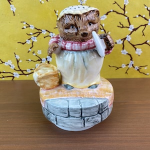 May include: A ceramic musical figurine of a hedgehog wearing a red and white checkered scarf and holding a plate. The figurine is standing on a stone base and has a basket in its hand.
