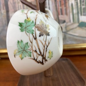 May include: A white porcelain teacup with a floral design featuring green and brown leaves and yellow accents. The cup is displayed on a wooden stand.