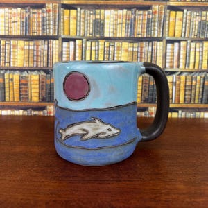 May include: A ceramic mug with a dark brown handle featuring a light blue upper section with a circular maroon sun and a darker blue lower section depicting a white dolphin in relief. The design is outlined with dark, etched lines throughout.