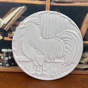 May include: A round, light pink ceramic coaster featuring a detailed relief of a rooster. The rooster is depicted in profile, with its tail feathers fanned out. The coaster has a slightly textured surface and a decorative border.