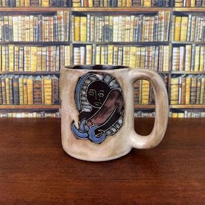 May include: A beige ceramic mug with a dark brown interior and a thick handle. The exterior features a hand-painted, raised relief design of a stylized face in black, surrounded by a crescent shape in shades of blue and a dark brown accent.