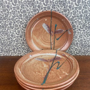 Può includere: Una pila di quattro piatti in ceramica fatti a mano. I piatti sono di colore marrone rossiccio macchiettato con un disegno dipinto di foglie e linee turchesi e verdi. Un piatto è impilato sopra gli altri.