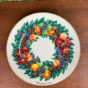 Könnte beinhalten: Ein weißer Zierteller mit goldenem Rand, der ein Kranzdesign aus grünen Blättern, roten Äpfeln, Pfirsichen, Trauben und Tannenzapfen zeigt. Der Text "Christmas 1988" ist unten aufgedruckt. Ein festlicher Teller zum Thema Feiertage.