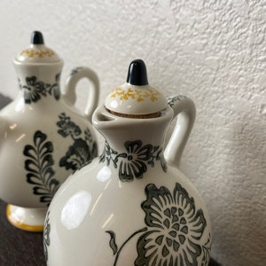 Może przedstawiać: Dwie białe ceramiczne butelki na olej i ocet z czarnymi i zielonymi wzorami kwiatowymi. Butelki mają korkowe korki z czarnymi wierzchołkami.