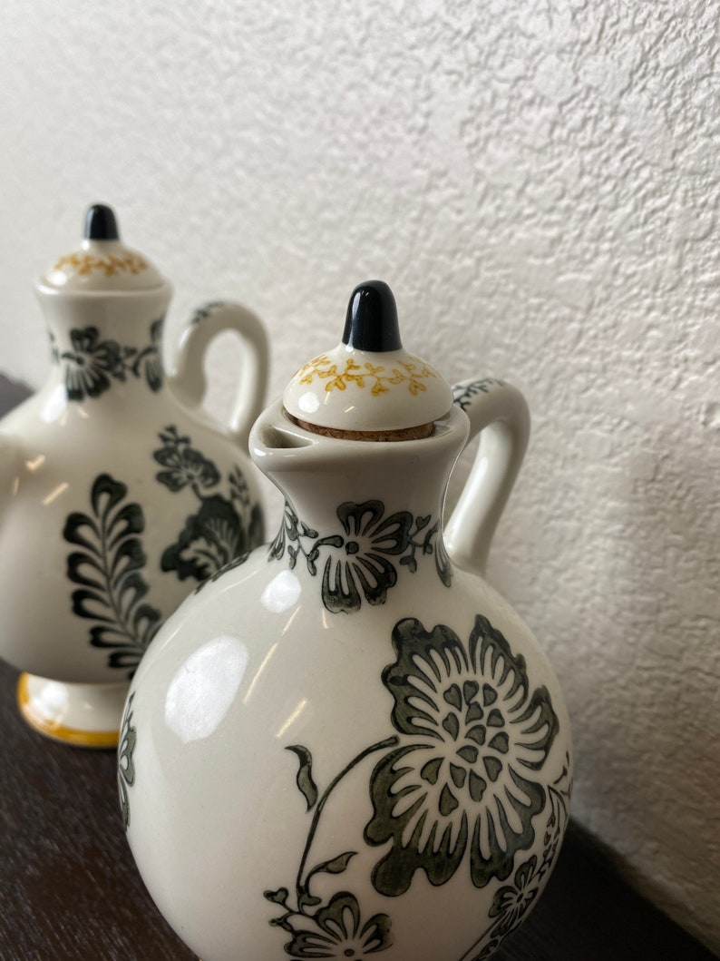 Może przedstawiać: Dwie białe ceramiczne butelki na olej i ocet z czarnymi i zielonymi wzorami kwiatowymi. Butelki mają korkowe korki z czarnymi wierzchołkami.