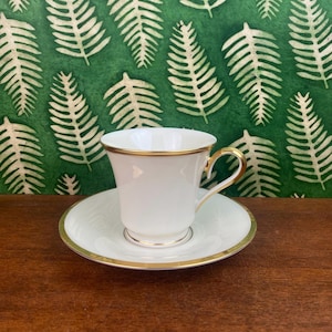 Könnte beinhalten: Ein weißes Tee-Set mit goldfarbenen Verzierungen. Die Tasse hat einen geschwungenen Henkel und steht auf einer passenden Untertasse. Der goldfarbene Rand verziert den Rand der Tasse und der Untertasse. Das Set steht auf einer Holzoberfläche.
