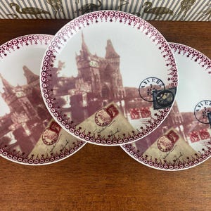May include: Three white ceramic plates with a burgundy border and a vintage image of a building with a red and white stamp that says "VIERS" on each plate.
