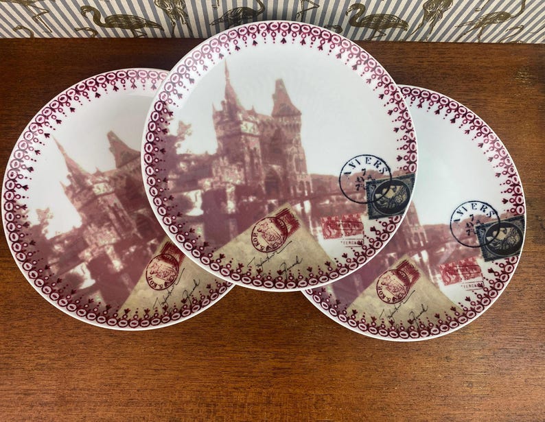 May include: Three white ceramic plates with a burgundy border and a vintage image of a building with a red and white stamp that says "VIERS" on each plate.