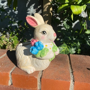 May include: White ceramic bunny planter with a pink nose and blue and pink flowers. The bunny is sitting on a brick wall.