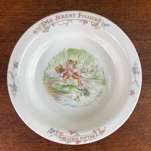 May include: White ceramic bowl featuring an illustration of Mr. Jeremy Fisher, a frog character, in a pond scene. The bowl has floral accents and the text "Mr Jeremy Fisher" and "Beatrix Potter" in banners.