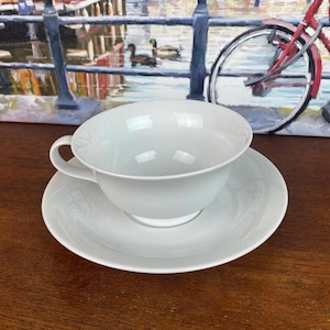 May include: A white teacup and saucer set. The cup has a rounded shape with a handle, and the saucer is circular. The set is made of ceramic and is placed on a wooden surface. The background includes a painting of a canal scene.