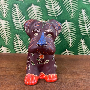 Può includere: Una statuetta di cane in ceramica dipinta a mano con dettagli in marrone, arancione, verde, blu e giallo. Il cane è seduto con le zampe in avanti e ha un motivo floreale sul corpo.