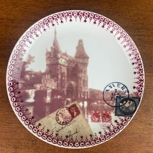 May include: A white ceramic plate with a burgundy border and a vintage image of a castle. The plate features several vintage postage stamps with the words "VIERS" and "TERRA" visible.