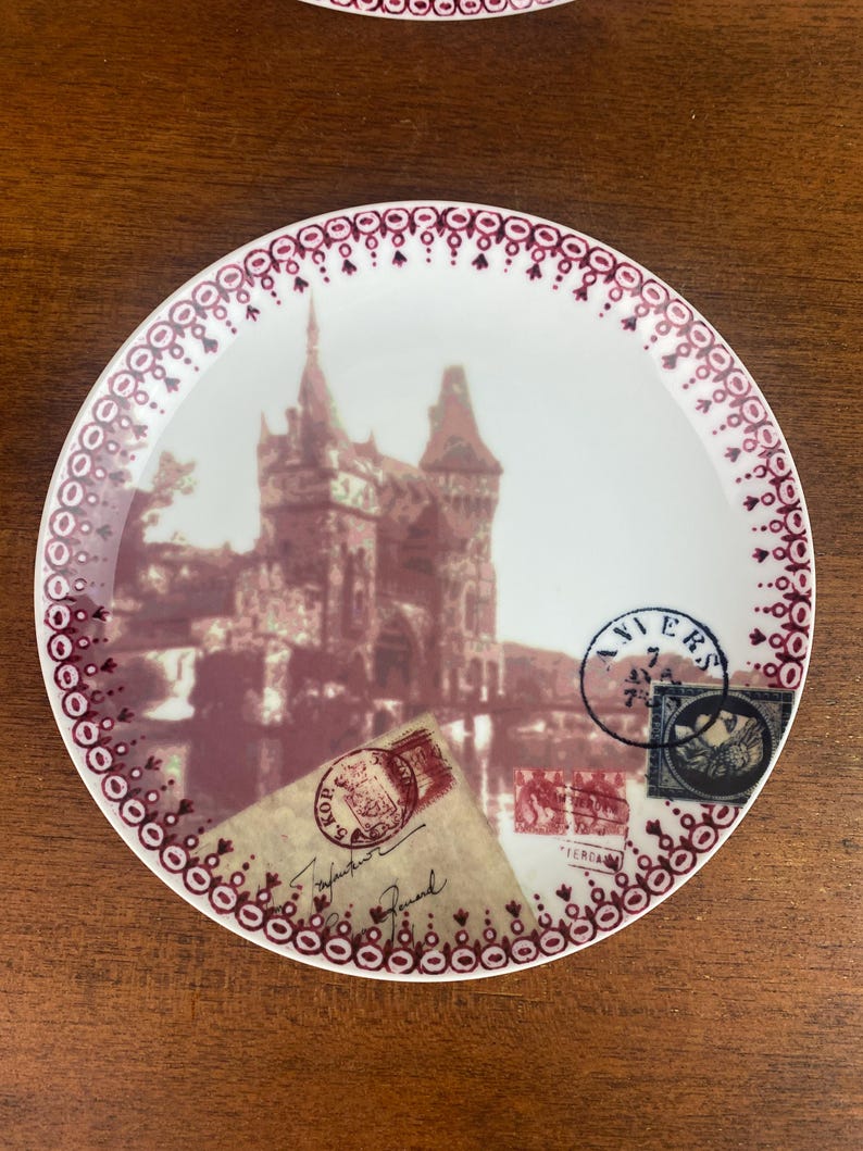 May include: A white ceramic plate with a burgundy border and a vintage image of a castle. The plate features several vintage postage stamps with the words "VIERS" and "TERRA" visible.