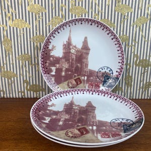 May include: Three white ceramic plates with a burgundy trim and a vintage image of a castle with a red roof and a brown background. The plates have a burgundy trim and a vintage image of a castle with a red roof and a brown background. The plates have a burgundy trim and a vintage image of a castle with a red roof and a brown background.